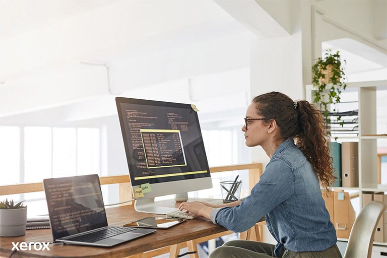 Eine Frau an ihrem Schreibtisch, beschäftigt mit einem Computer und einem Laptop, demonstriert Produktivität an einem modernen Arbeitsplatz.